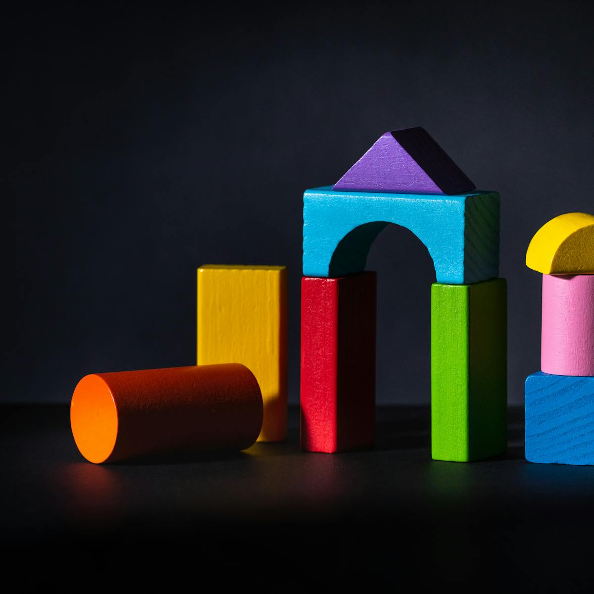 A photograph of multi coloured wooden blocks against a black background. The blocks are of various shapes and stacked on top of each other. The blocks appear so that half of the block is in dark shadow and silhouette.
