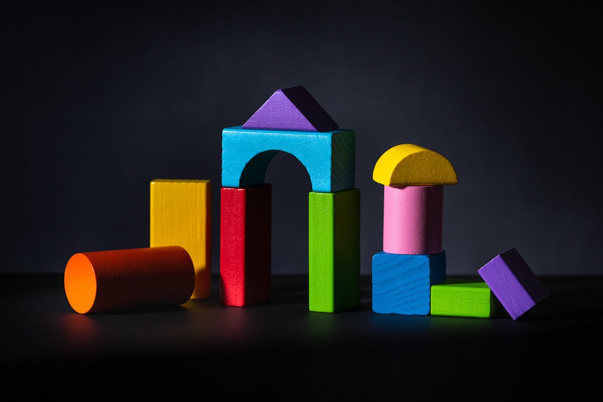 A photograph of multi coloured wooden blocks against a black background. The blocks are of various shapes and stacked on top of each other. The blocks appear so that half of the block is in dark shadow and silhouette.
