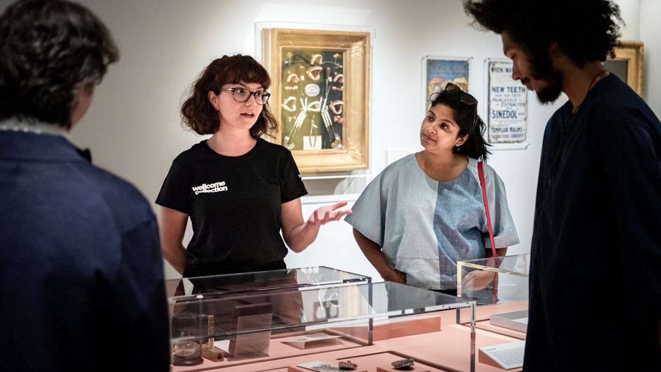 Photograph of a young female museum gallery visitor experience assistant wearing a black T-shirt with 'Wellcome Collection' written on it, giving a tour of the exhibition space. She is stood behind a glass waist high glass display case containing objects, her left hand raised in explanation. To her left and right are members of the public listening to her speak. Behind her on the gallery wall are posters and framed exhibits.