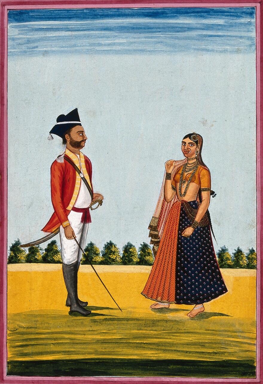 Gouache painting of a sepoy, a soldier, and a woman standing outdoors. He is wearing traditional military dress - a red coat with yellow trim, a red sash around his waist, white trousers and a black hat. He is wearing long black boots and a sword which is in a scabbard. He is holding the sword and it is trailing on the ground. The woman is standing to the right and is wearing an orange cropped blouse and a long patterned orange and navy skirt. She is wearing a lot of elaborate jewellery. Behind the couple is a row of trees in the distance, the sky is blue. 