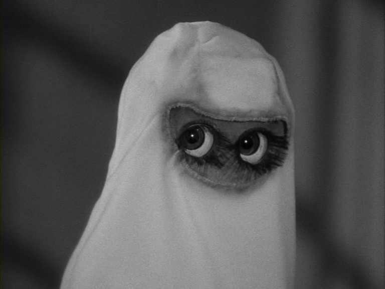 An alien creature with a blanket over its head looks at the viewer through a hole cut for its eyes.