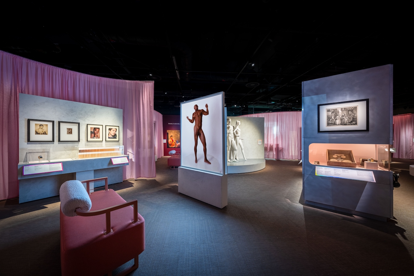 An exhibition space with several display areas showing images and objects on vertical sections of wall. In the foreground is a sofa infront of a standalone screen projecting a video of a male figure in motion.