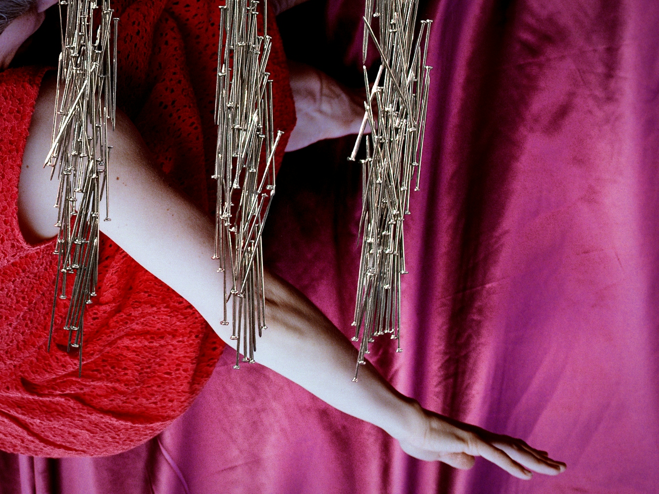 Detail from a larger artwork created with a colour photographic print of a female figure in a bright red dress, set against a purple and blue draped silk background. The detail shows the woman's outstretched arm and three of the pillars made out of dress pins.