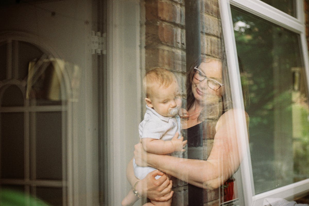 Photograph of a mother holding her baby son taken through the glass of a window. The two are looking down, their heads slightly bowed. The mother has a smile on her face and the baby has a dummy in his mouth. The scene around them is fairly abstract and dream-like due to the reflections, but part of an open front door can be seen, along with brickwork and green foliage. 