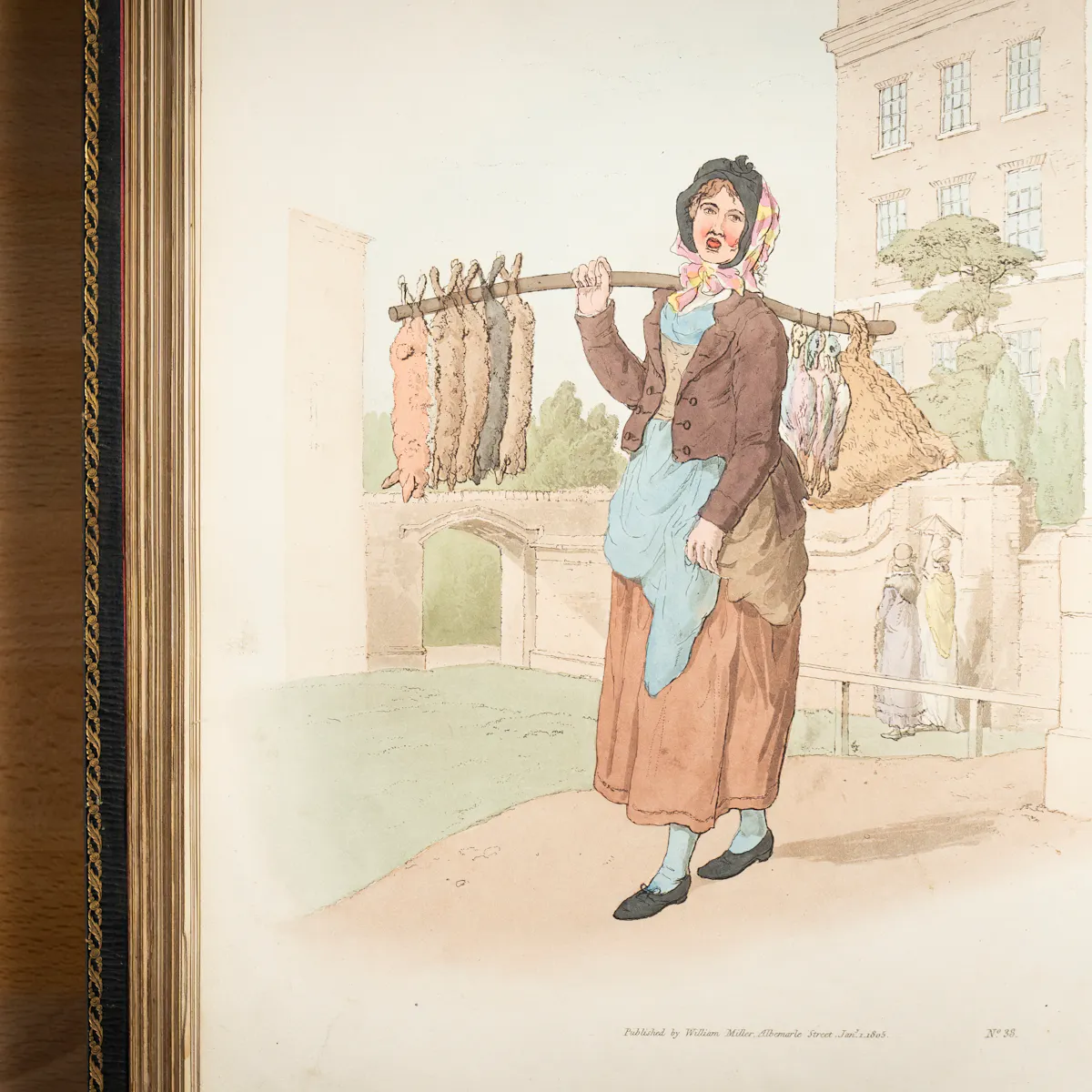 A photograph of a book containing colour illustrations. The illustration depicts a working class woman with her mouth open and carrying a number of dead rabbits arranged along a stick which she holds over her shoulder. In the background are upper class ladies walking along under their parasol.