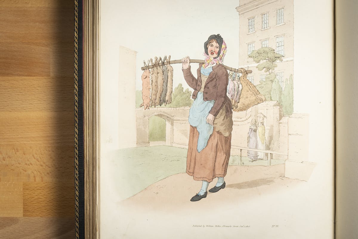 A photograph of a book containing colour illustrations. The illustration depicts a working class woman with her mouth open and carrying a number of dead rabbits arranged along a stick which she holds over her shoulder. In the background are upper class ladies walking along under their parasol.