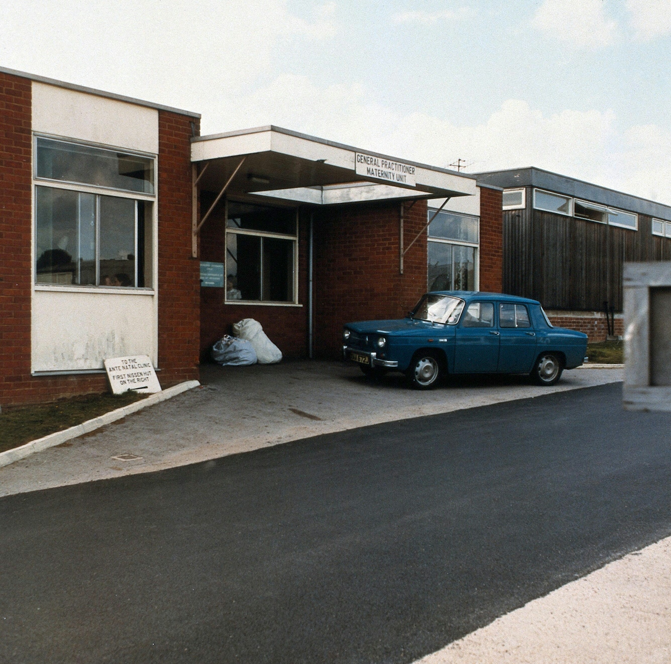 Colour photograph of the outside of the maternity unit at the Churchill Hospital, Oxford, in 1972. A small blue car is parked outside the brick building and bags of laundry lay under the front awning, which bears a sign reading "General practitioner maternity unit". A sign leaning against the bottom of a window reads "To the antenatal clinic, first Nissen hut on the right".