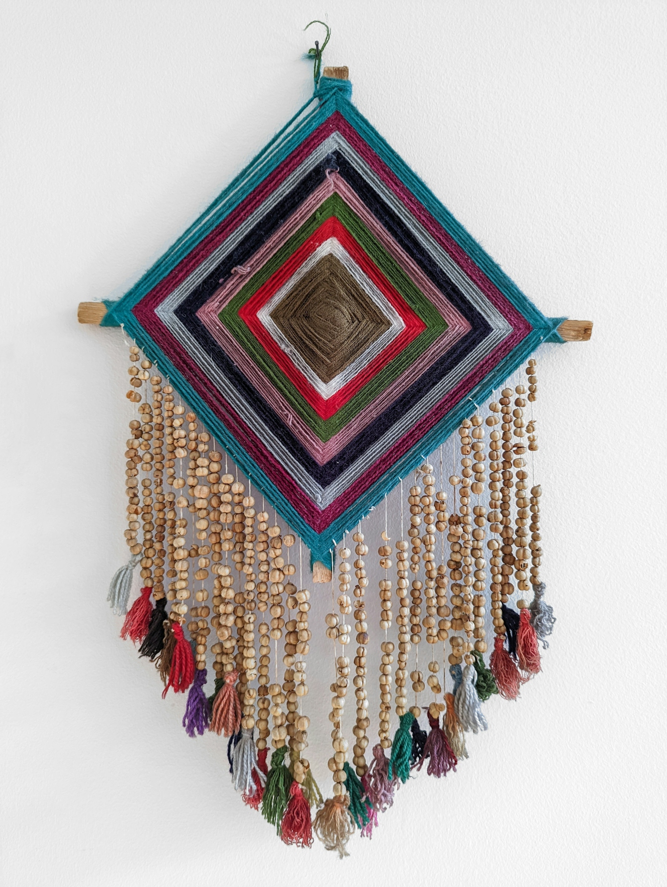 Wall-hanging made of different coloured strands of wool woven in stripes into a diamond shape on a wooden frame. Strands of rue seeds threaded onto string hang from with bottom edge of the diamond, each ending in a woolen tassle.