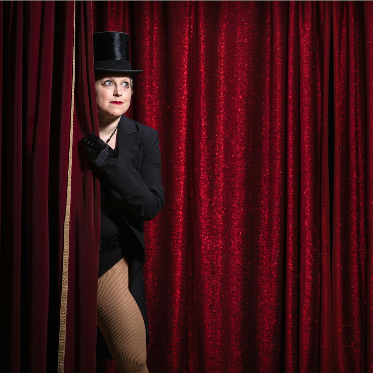 Photograph of a woman dressed in a top hat and tails peeping around the red curtain on a theatre stage. Behind her is another glittering red curtain.