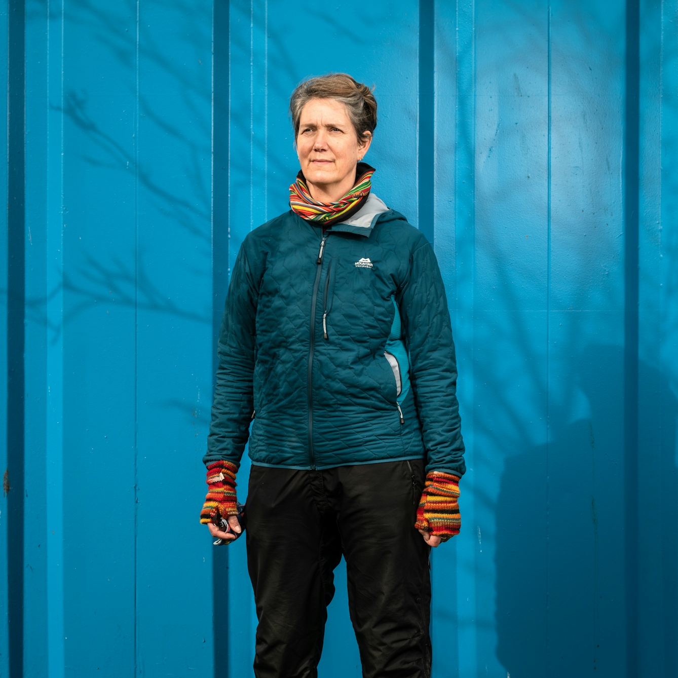Photographic portrait of a woman from the knees up, wearing a blue insulated jacket, a colourful stripy scarf and colourful stripy fingerless gloves. She is stood against a blue shipping container and the shadow of a leafless tress is cast across her and the background, by the sun. She is looking off into the distance to the left of the camera.