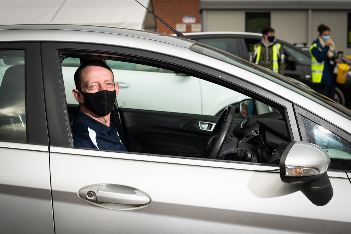 Photographic flash lit portrait through the open side window of a stationary car. The car is silver and the front of the car is facing to the right. Sat in the driver's seat is a man, looking to the camera. He is wearing a black face covering. In the background is the out of focus side of a white marquee, distant buildings and 2 people wearing hu-viz vests.