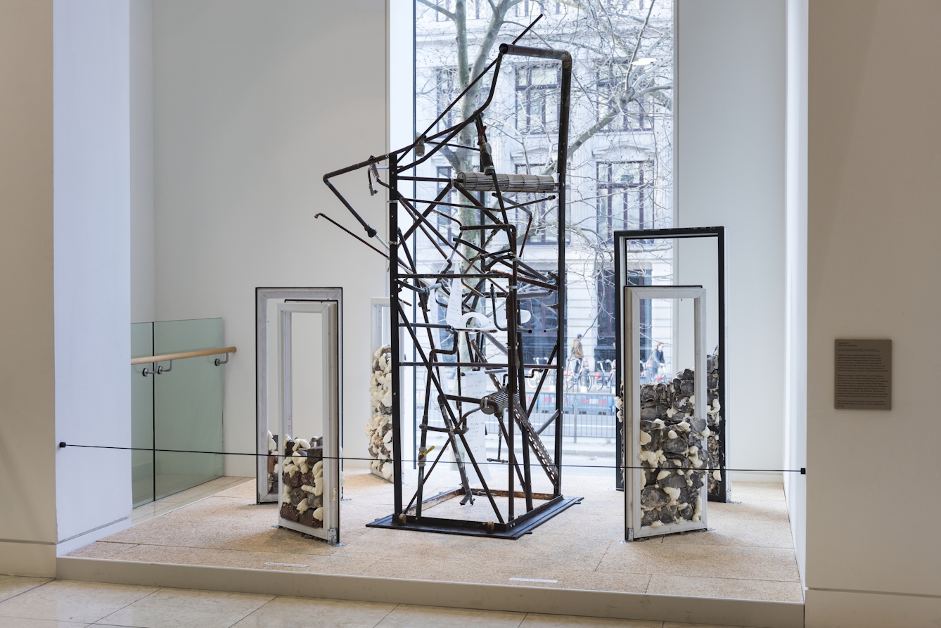 A photograph of an artistic installation posiitoned in a window, consisting of a rectangular cuboid metal structure in the centre surrounded by five UPVC-window-like structures filled with rocks and expanding foam. 