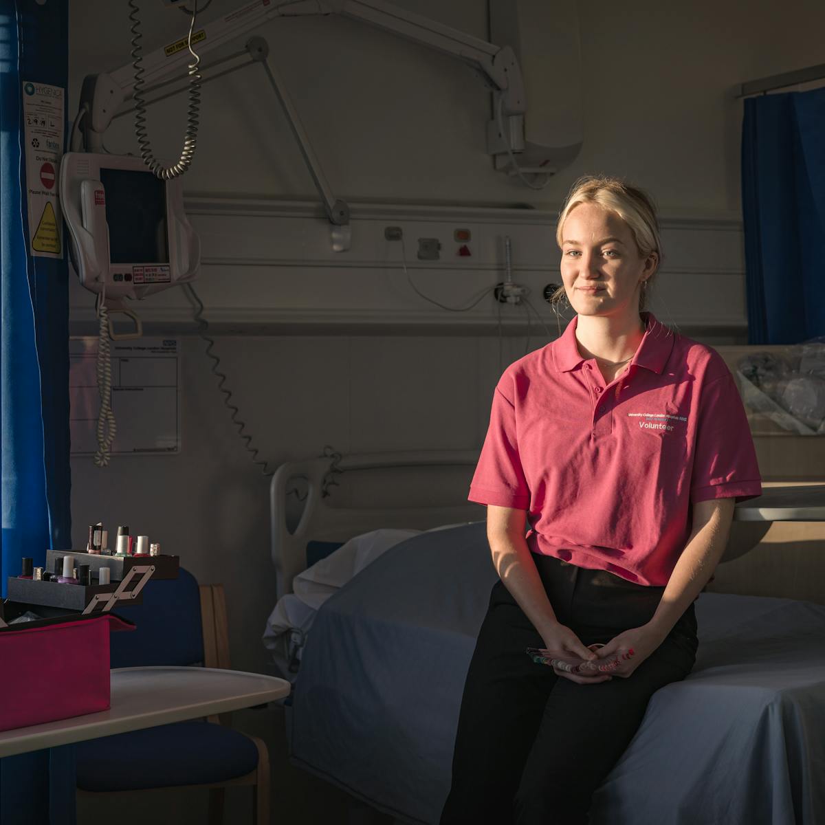 Photograph of a young woman sitting on the side of a bed in a hospital ward, light by the light of the setting sun streaming in through the window. She is wearing a pink polo shirt smiling to camera. Behind her are the blue bay curtains and monitoring devices of a typical hospital setting. On a table to the left is an open pink case containing nail varnish beauty products.