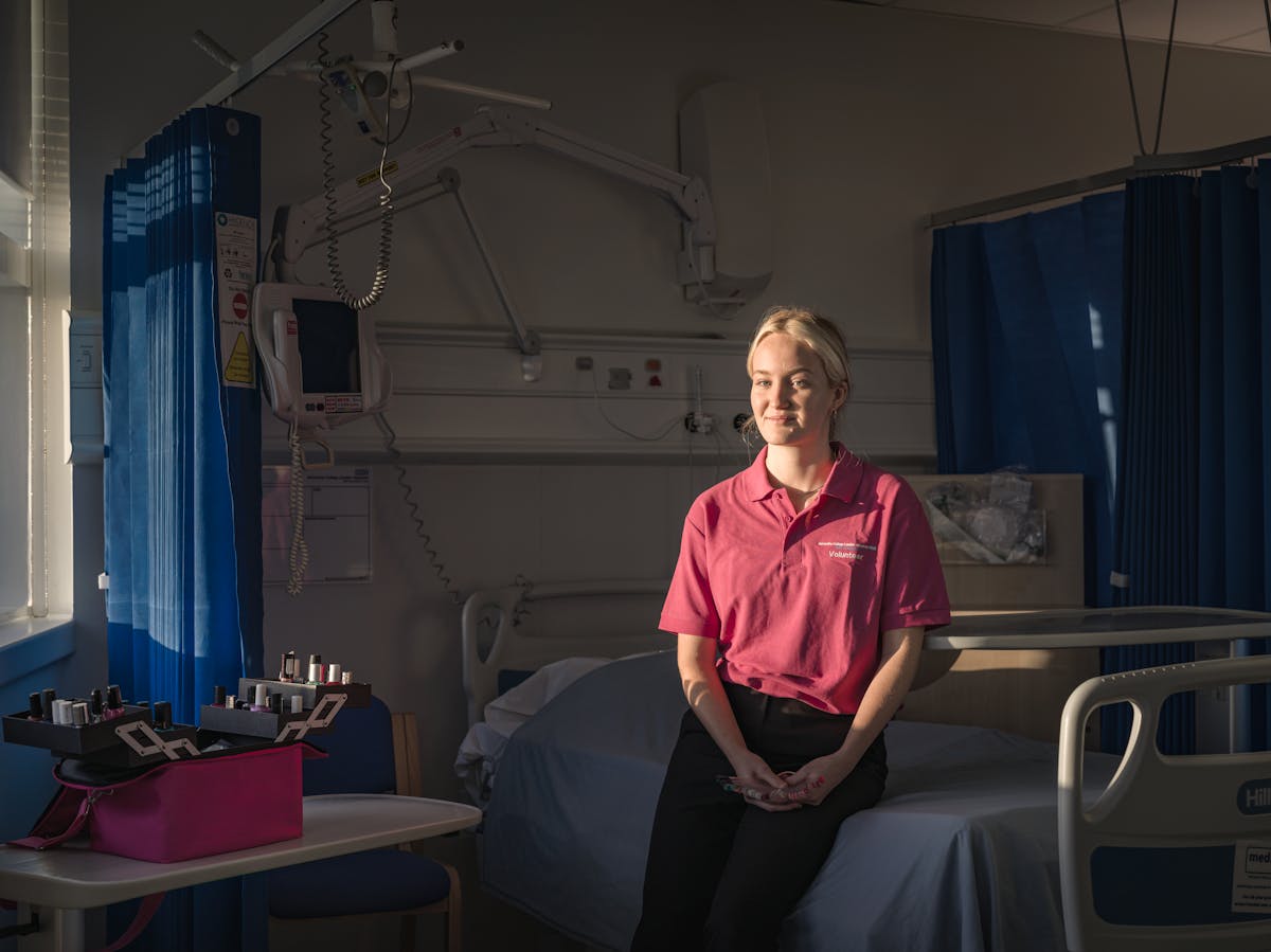 Photograph of a young woman sitting on the side of a bed in a hospital ward, light by the light of the setting sun streaming in through the window. She is wearing a pink polo shirt smiling to camera. Behind her are the blue bay curtains and monitoring devices of a typical hospital setting. On a table to the left is an open pink case containing nail varnish beauty products.