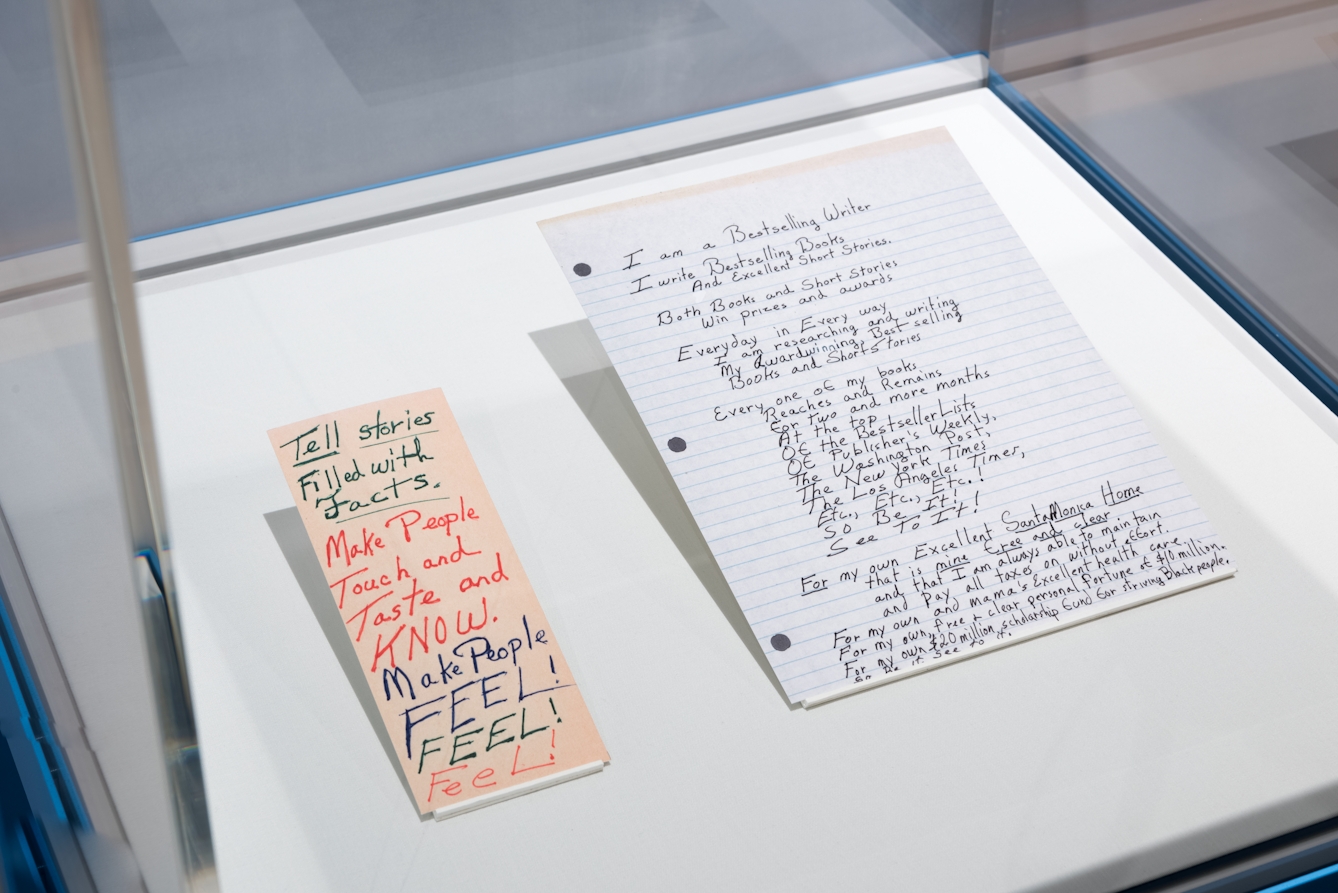 Photograph of an exhibition glass display case showing two paper exhibits raised at an angle off the display case base. The paper on the left is orange in colour and long and thin. On it are written the words, 'Tell stories filled with facts. Make people touch and taste and KNOW. Make people FEEL! FEEL! FEEL! The paper on the right is lined and has 3 hole punches. It contains a full page of text which starts, 'I am a Bestselling Writer...'