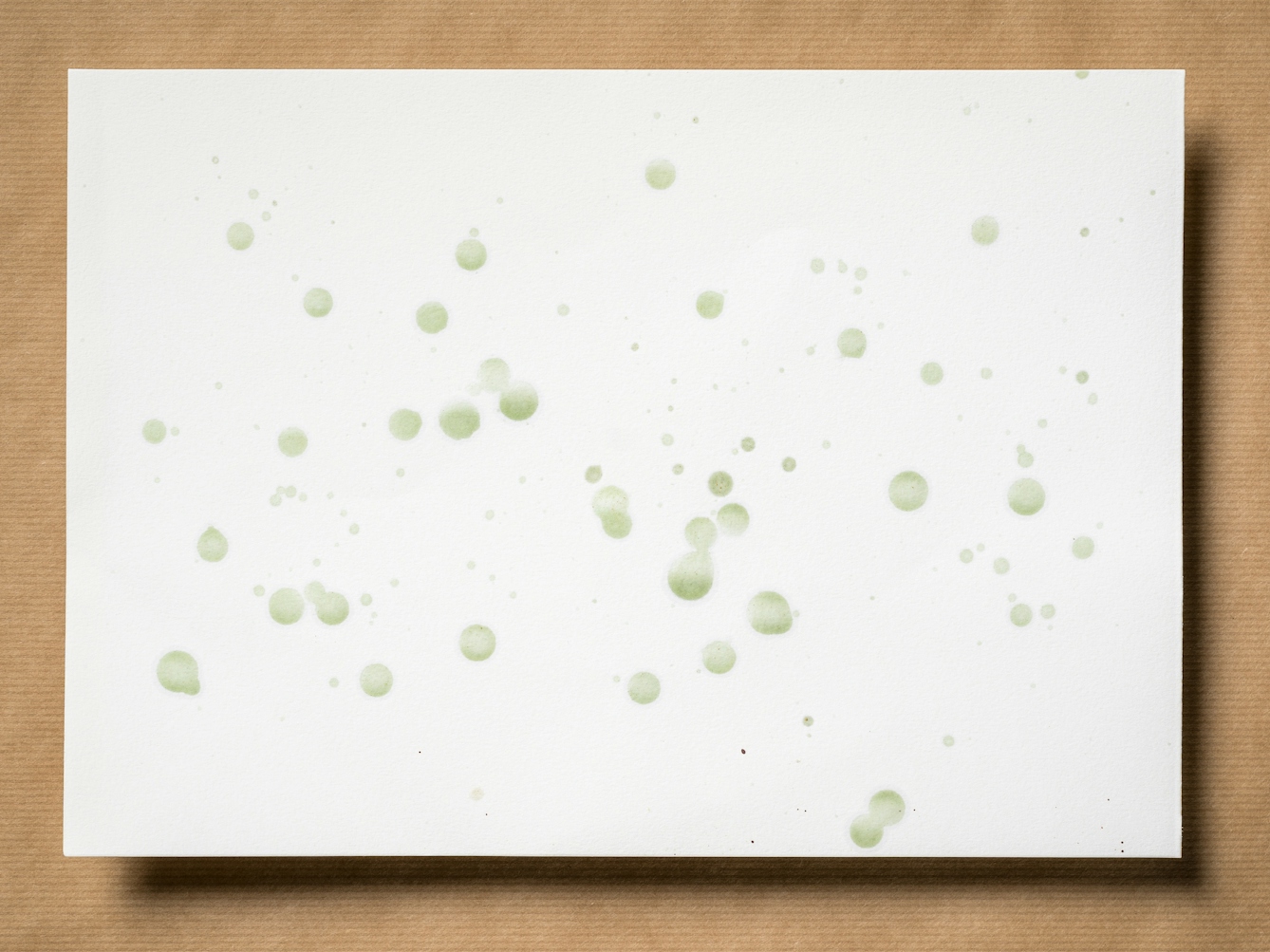 Photograph of an original artwork on watercolour paper. The artwork is resting on a brown parcel paper background. The artwork shows many varying in size droplet spatters made with coloured ink. The ink colours are green. The spatters seem random in distribution.