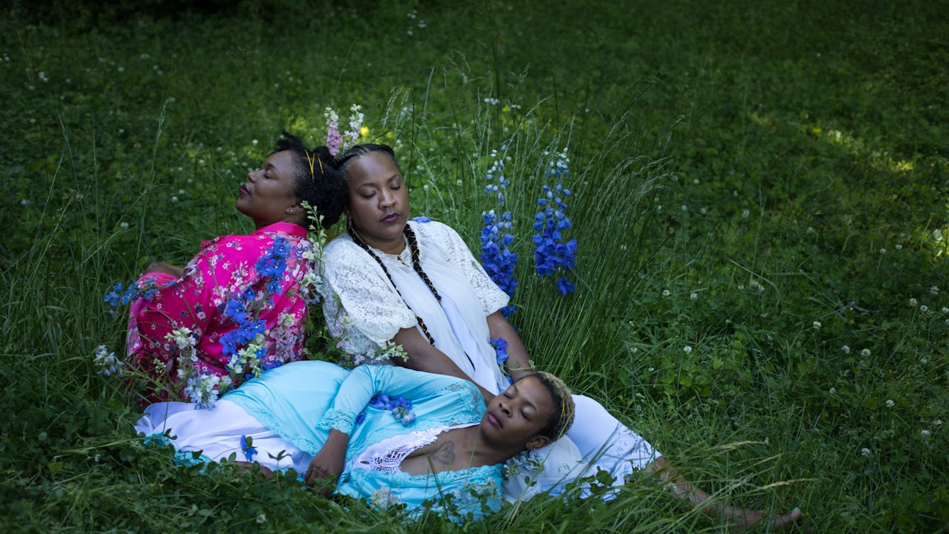 Portrait of Tricia Hersey in a field with her eyes closed leaning on two women. They are sleeping in nightwear among tall grass and flowers.
