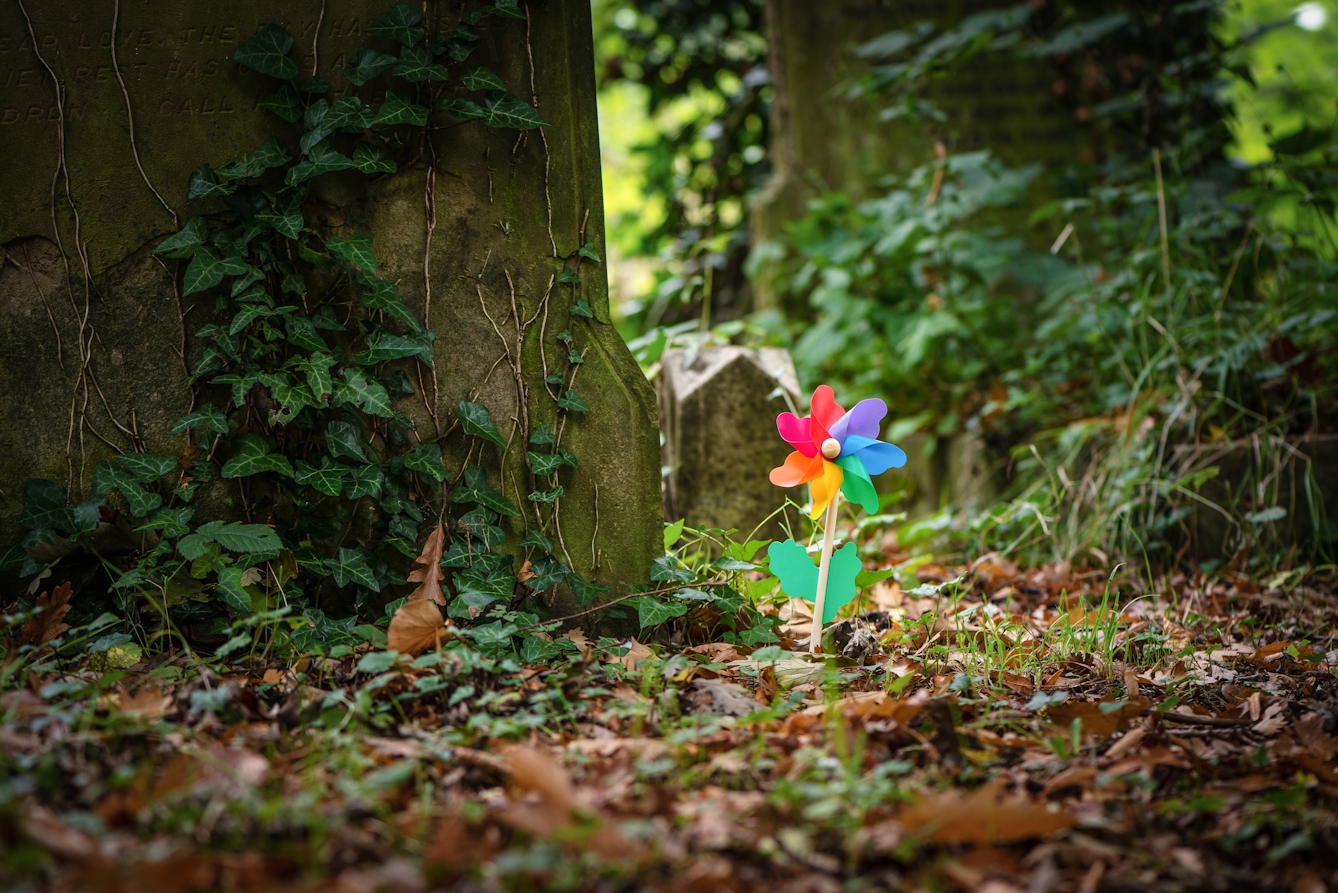 Photograph of a colourful windmill sitting within the surroundings of a cemetery, with grave stones and ivy.