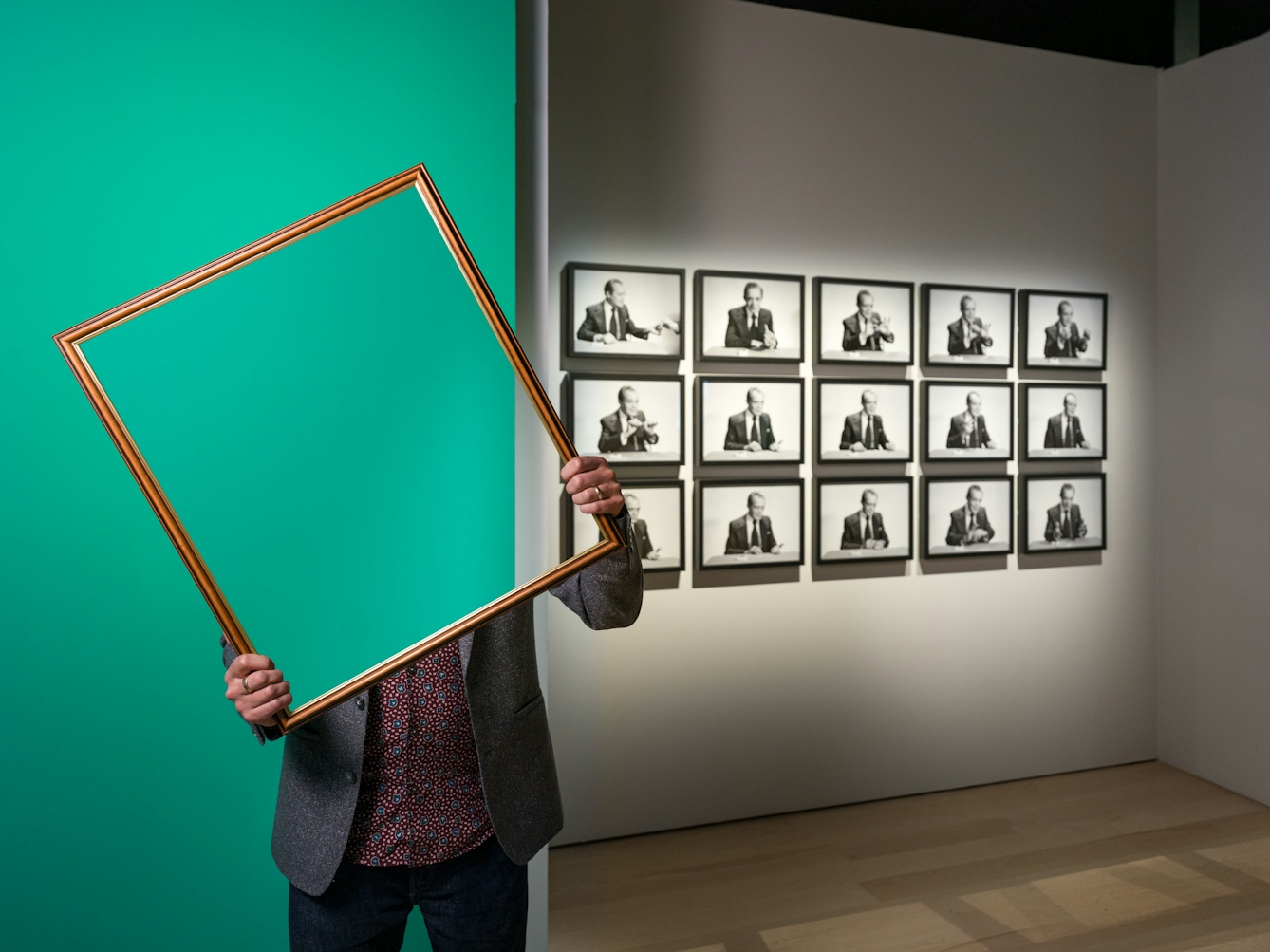 Photograph of a man in a gallery space holding a picture frame in front of his face and upper body. Within the picture frame you can see straight through him, as if his body and head have disappeared. In the background you can see a green painted wall and a grid of framed black and white photographs.