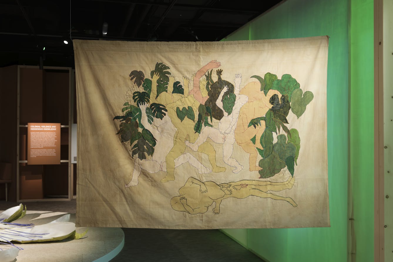 Photograph of an artwork embroidered in green and earthy tones on a large sheet, depicting parts of humans and parts of plants. 