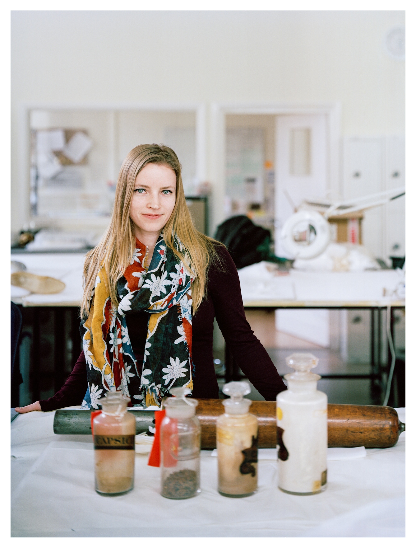 Photographic portrait of a woman standing behind a table looking to camera wearing a black top and colourful floral pattered scarf. Behind her is a museum conservation environment with tables and lights and objects. Visible on the table in front of her are 4 large glass bottles with stoppers and a large wooden object laid on its side.