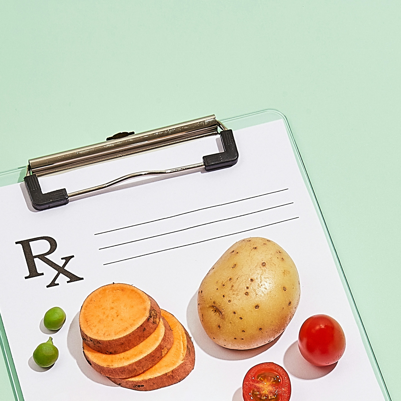 Photograph of a clipboard with a blank doctor's prescription note attached to it, resting on a light green background. On top of the paper prescription are a whole potato, a stack of sliced sweet potato, whole and halved cherry tomatoes and green peas.
