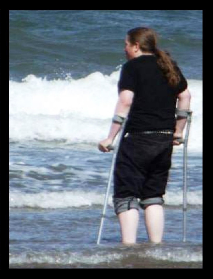 A person standing on a beach looking out at the waves with their feet in the sea to their ankles. Their jeans are rolled up to the knees and they are holding an elbow crutch in each arm