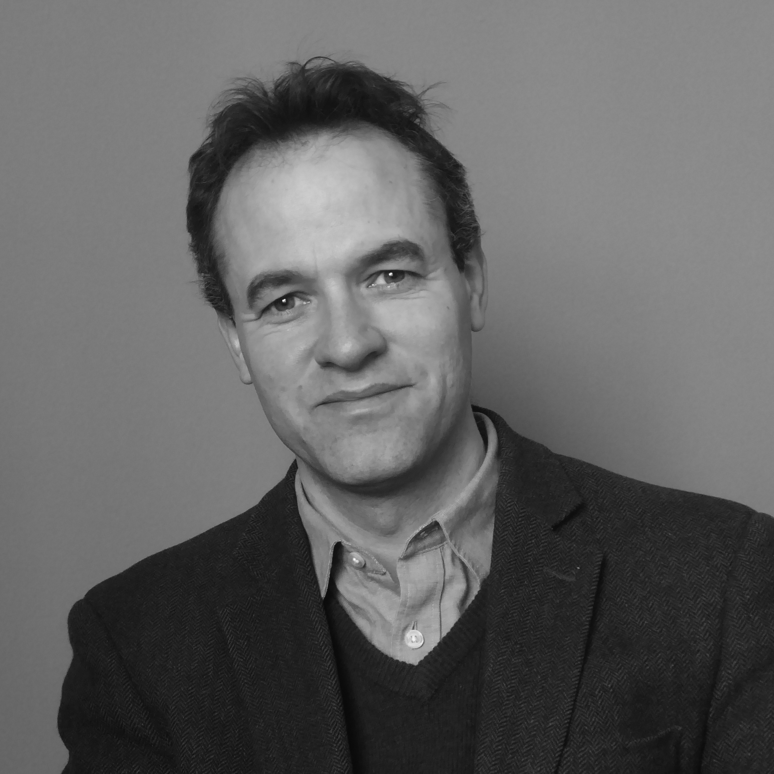Head and shoulders photograph of a man with short hair, wearing a shirt and blazer standing in front of a grey background.