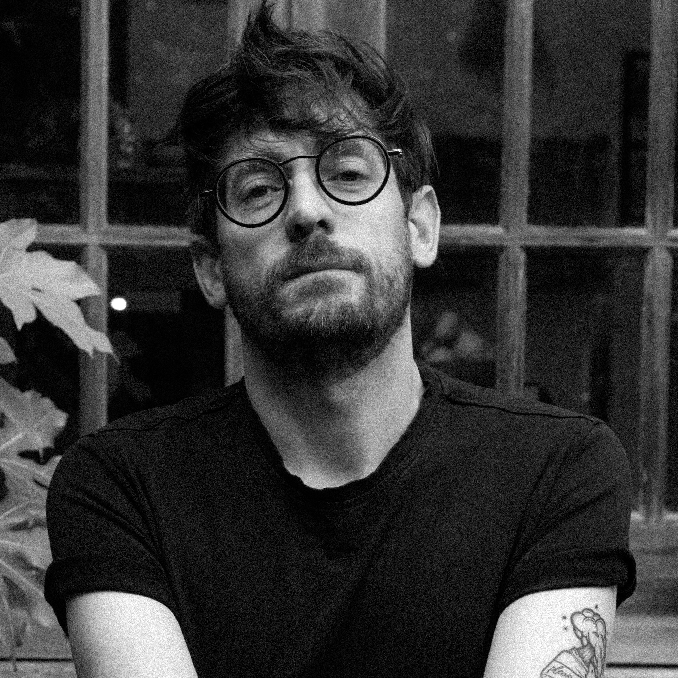 Black-and-white headshot of a person with short hair, round glasses and a beard, wearing a dark T-shirt.