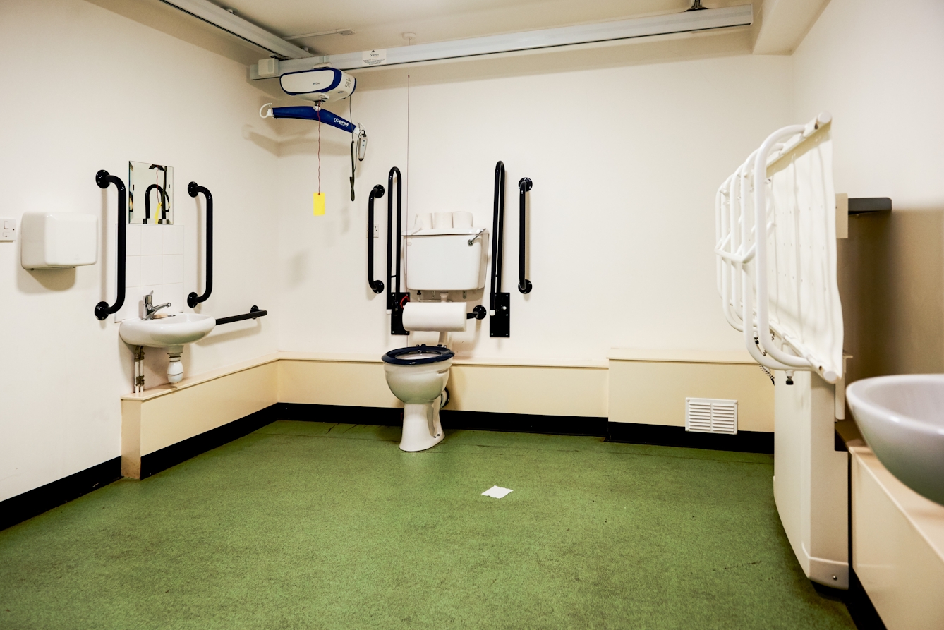 Photograph of the interior of an accessible toilet showing a green floor and beige walls. Attached to the ceiling is a rail mounted hoist and on the right hand wall is a large fold down changing table.