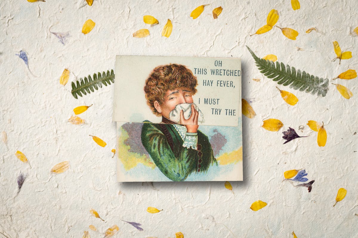 An image of 19th century advertising card for medicine, the card shows a woman with brown, wavy hair in a  green dress holding a handkerchief to her nose, with an expression of suffering on her face. Behind the card is a background of yellow, blue and violet petals as well as green leaves.