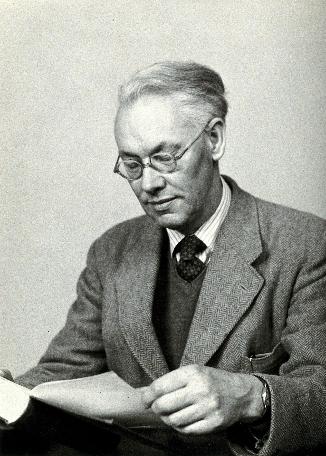 Black and white photograph of a middle-aged white man, Sir Christopher Howard Andrewes, wearing glasses and a tweed suit who is reading a book. 