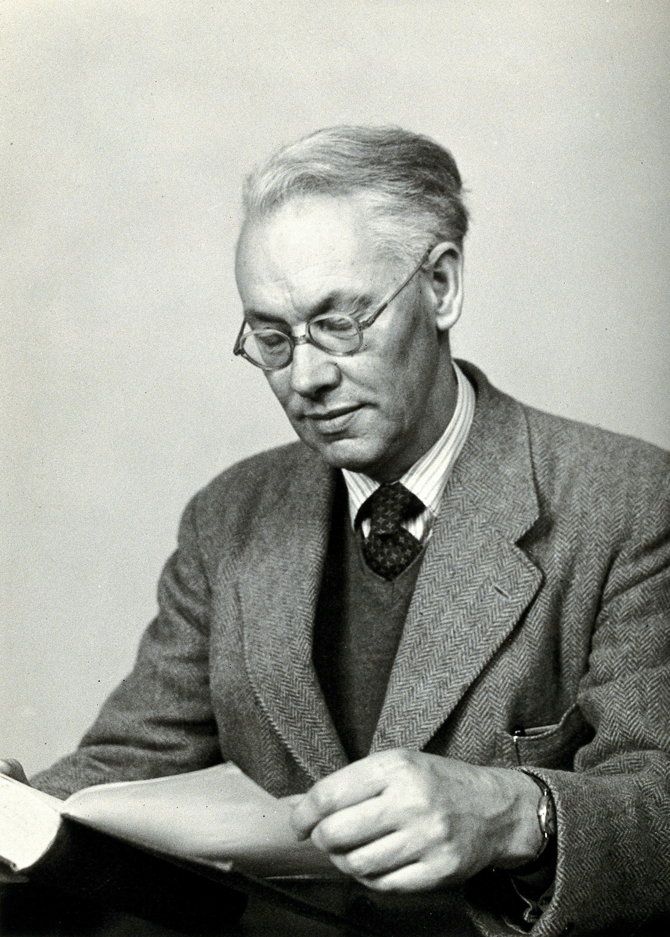 Black and white photograph of a middle-aged white man, Sir Christopher Howard Andrewes, wearing glasses and a tweed suit who is reading a book.