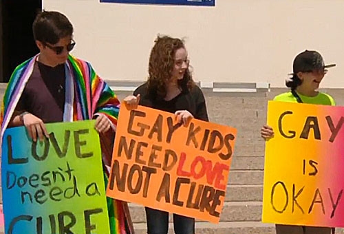 Teenagers hold signs saying 'Gay kids need love not a cure' and 'Gay is Okay'.