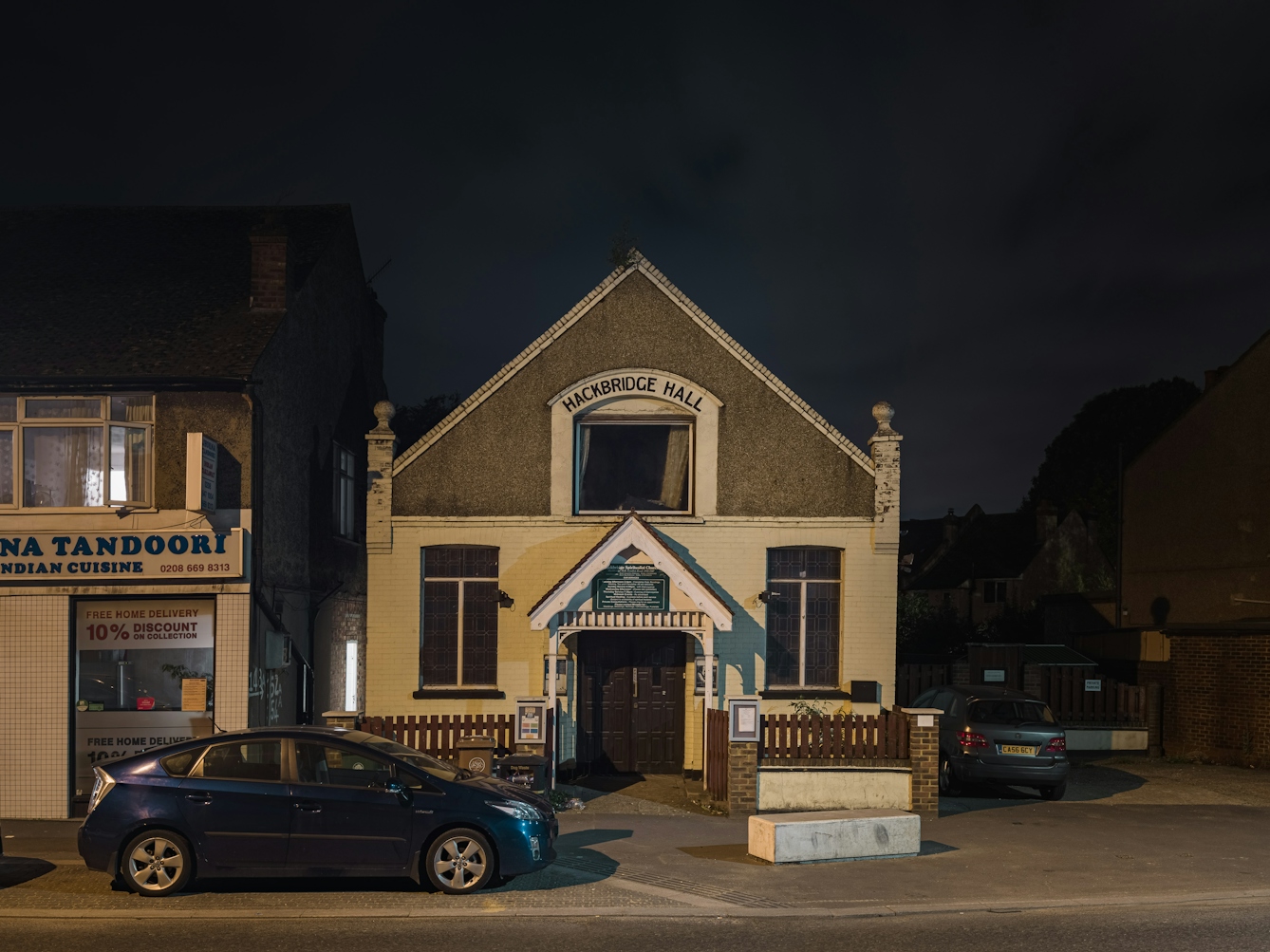 Photograph of Hackbridge Christian Spiritualist Church at night.  The double storey building is located in the centre of the frame, with a Indian cuisine restaurant to the left. of the building.  In the foreground is a blue sedan.