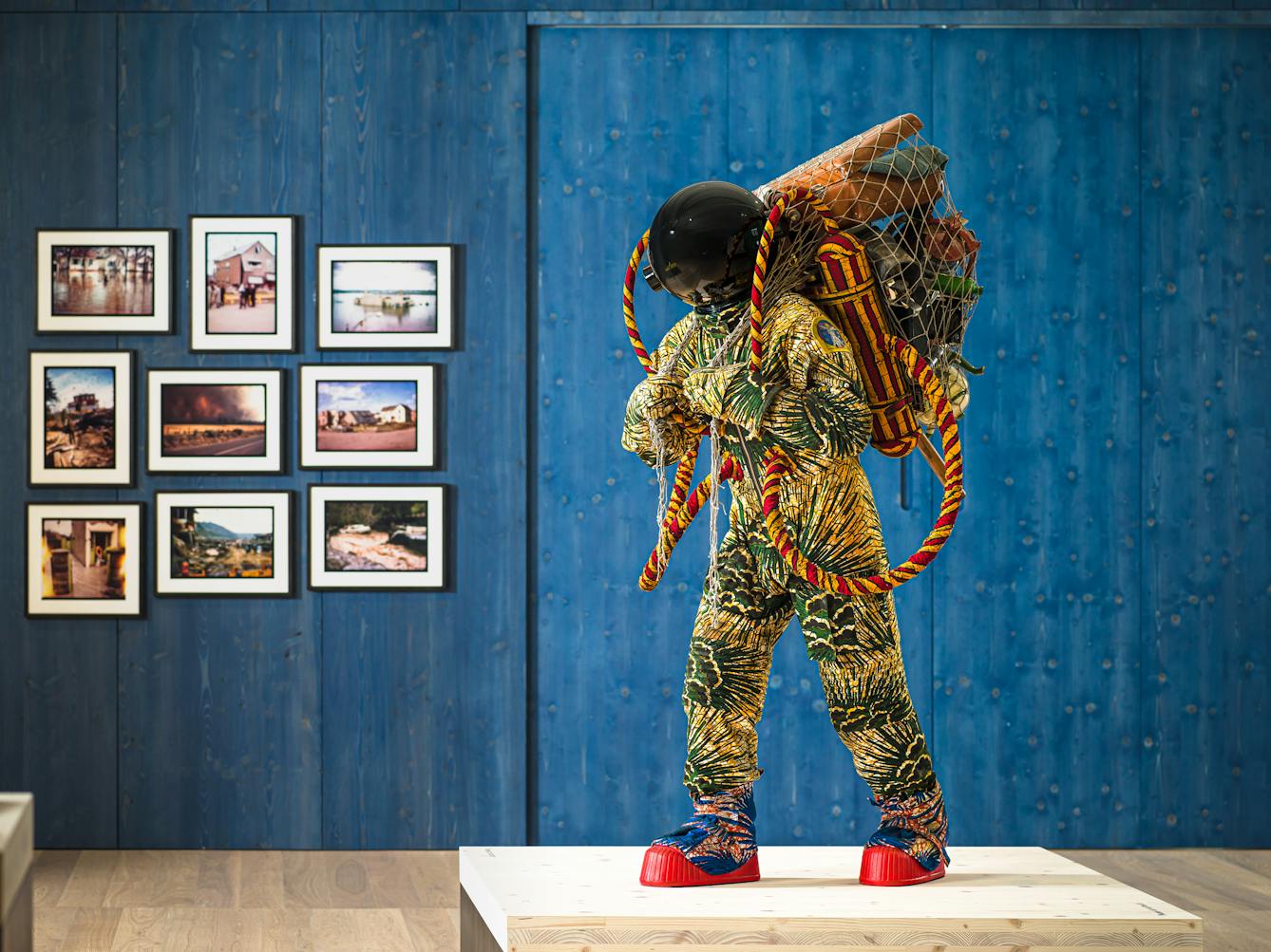 Photograph of an exhibition gallery space, with a blue stained wood wall in the background. In the foreground is a life-size artwork of a figure resembling an astronaut. carrying a large net containing assorted objects including a suitcase.