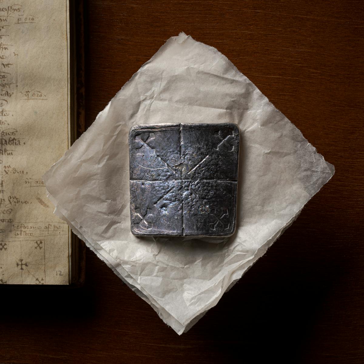 Photograph of a curved lead healing plate about ten centimetres square, sitting flat on a piece of conservation paper.  Below the lead plate, to the left, is the manuscript from which the original instructions to make the plate can be found.  The lead plate has been inscribed with 5 crosses; one diagonal cross in each corner with lines leading to a central, right-way-up, cross.