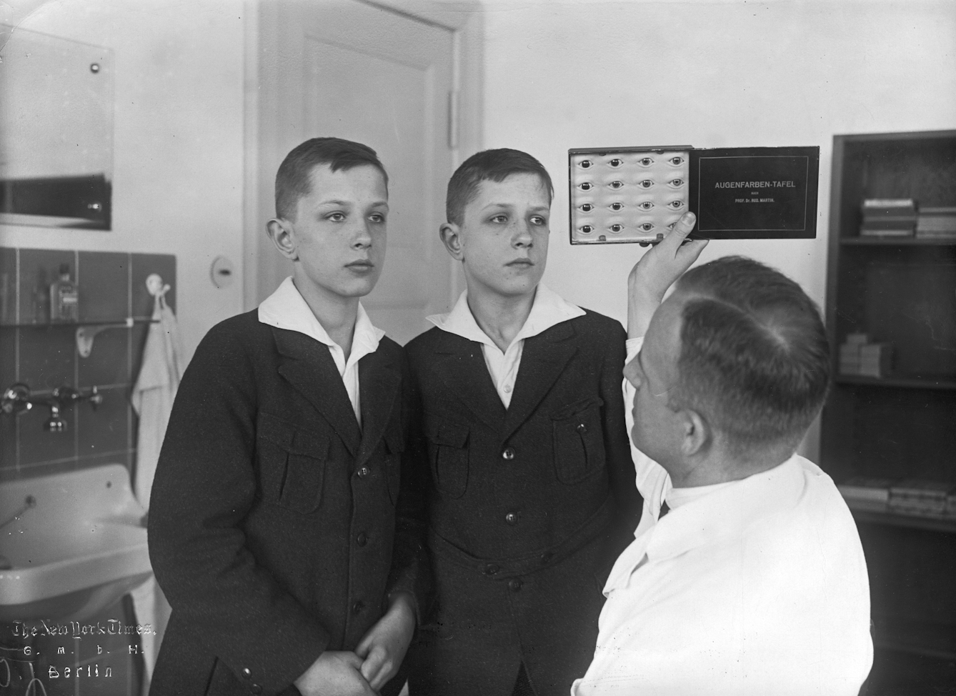 Black and white photograph of two adolescent, identical twin boys standing side by side in front of a man with a white coat who holds up a chart with illustrations of different eyes while looking at the eyes of the boys. The three figures are in a laboratory.