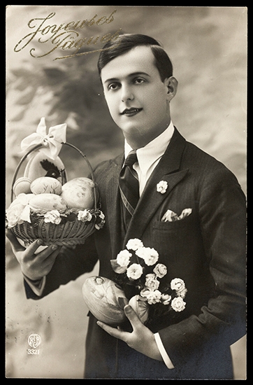 A young man in a smart suit with a basket of decorated Easter eggs in one hand and a single decorated egg and a bunch of flowers in the other hand.