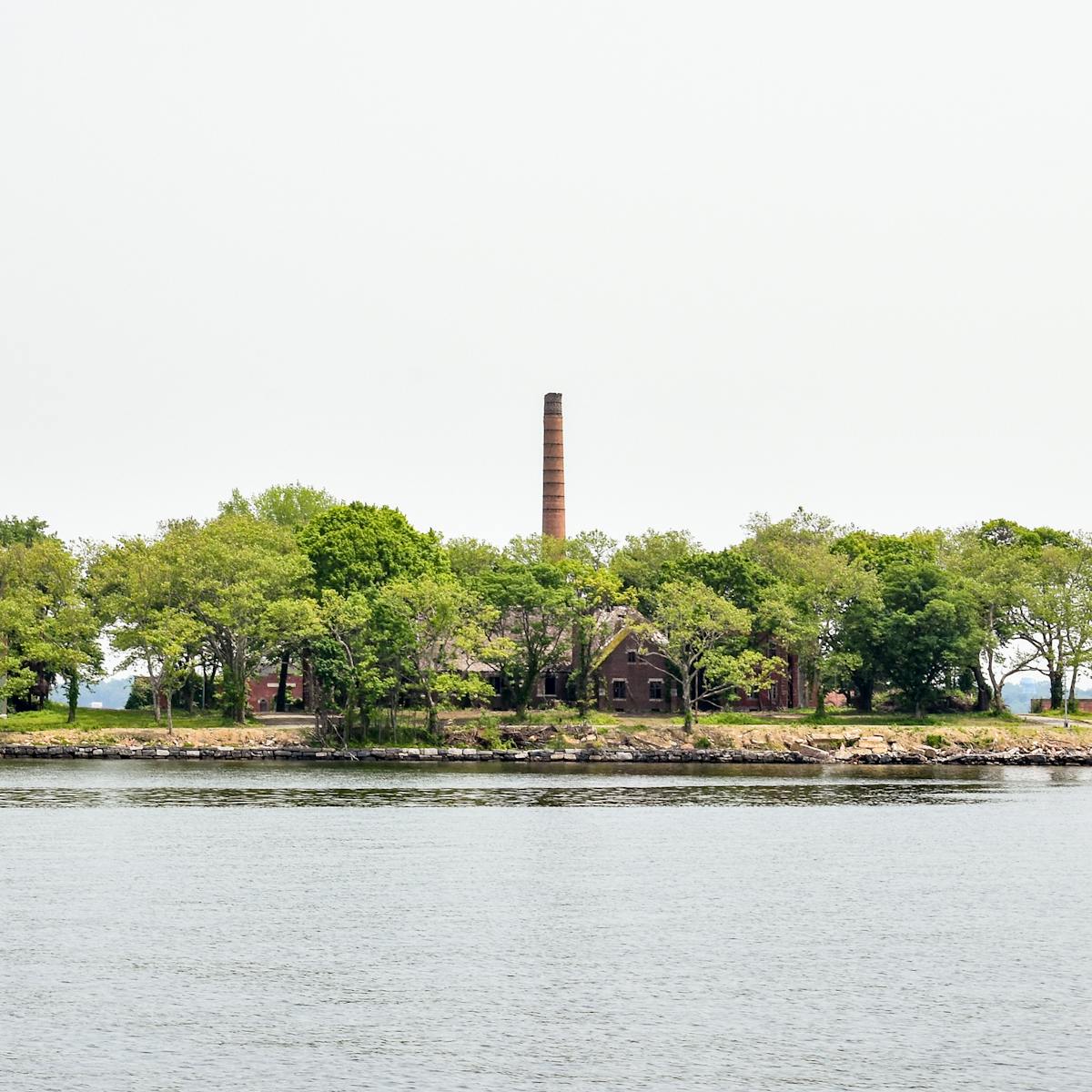 Photograph of a small island. The lower half of the image is water, the upper half and overcast sky. Across the centre of the image is the island, covered in green trees and several 2 and 3 storey buildings and a tall chimney in the centre.