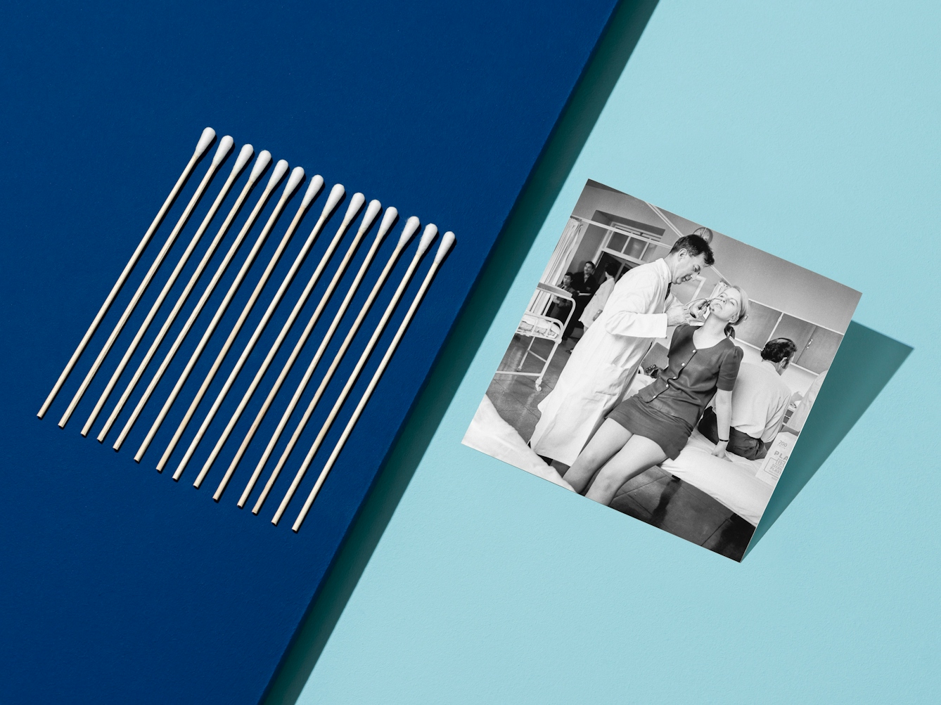 Photograph of clinical light blue background with a light blue surface floating above the dark blue and appearing from the left hand side, covering half the image. Propped up on the light blue surface is a black and white archive photograph showing a woman perched on the edge of a hospital bed whilst a male doctor administers an anti-flu vaccine up her nose. On the left hand side of the image is a row of neatly lined up medical swabs.
