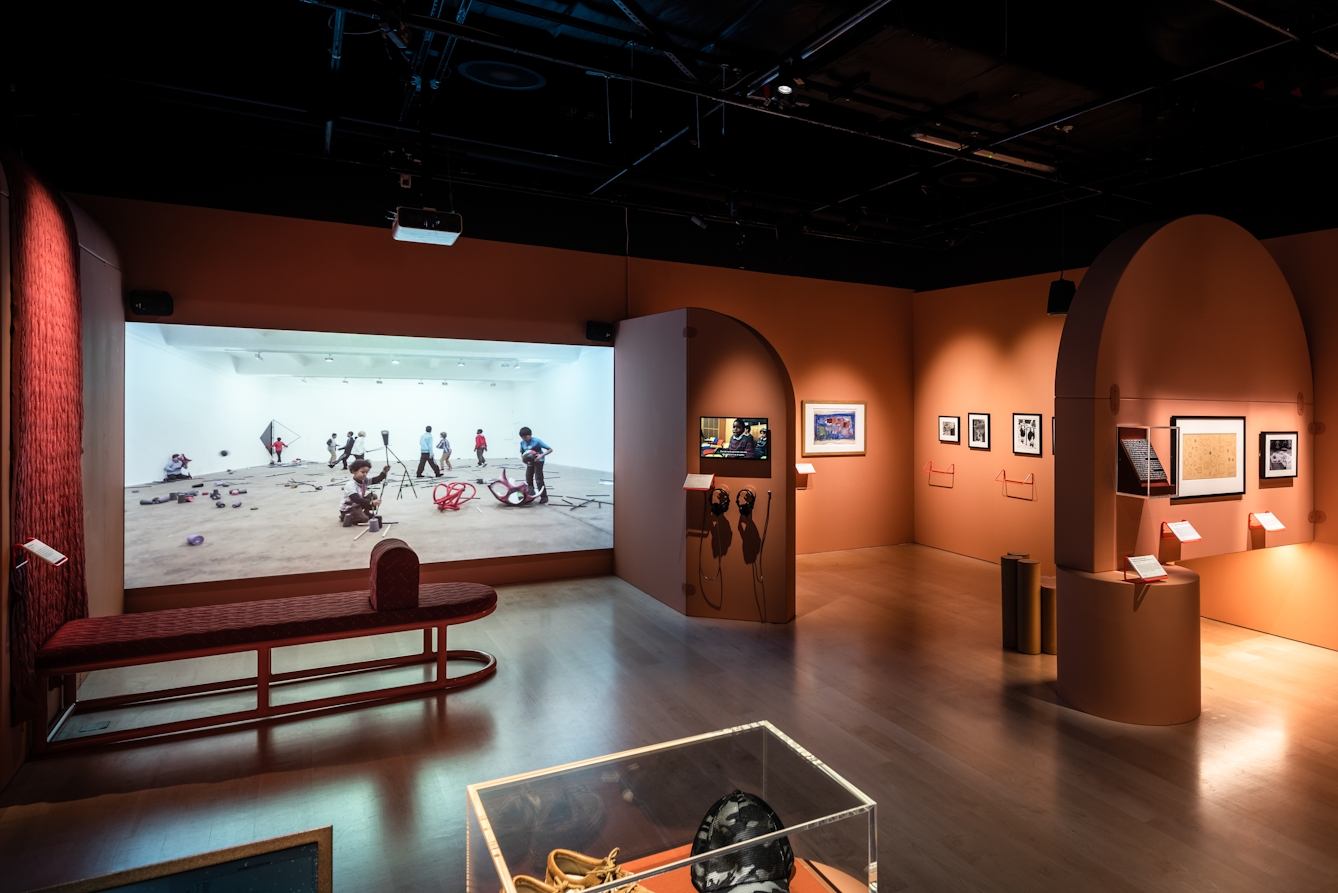Photograph of an exhibition gallery space containing orange walls with arched details and framed wall mounted exhibits. To the right of the frame is a projected film. The current film still shows young boys in a large white space surrounded by fragments of what were larger sculptures.