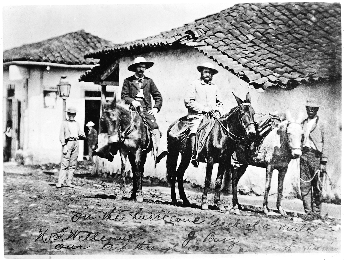 Black and white photograph of two European men on mules accompanied by a local man on foot standing in front of small buildings with tiled roofs in Ecuador in 1878