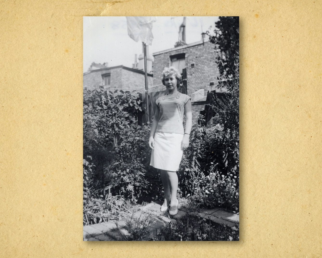 Photograph of a black and white photographic print, resting on a brown paper textured background. The print shows a woman in full length standing in the back garden of a row of houses. She is looking to camera, wearing a white skirt and stripped top. She is surrounded by shrubs and trees. Behind her are brick houses.