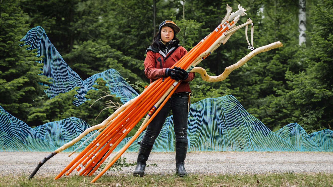 Photograph of a woman standing on a gravel path with a fir tree wood in the background. She is wearing outdoor cloths, gloves, wellington boots and a cap. Cradled in her arm are long orange sticks, some with animal jaw bones on the end. Interwoven into the scene is a graphic element made up of thin blue drawn lines, which create a mountain range behind the woman which disappears into the trees.