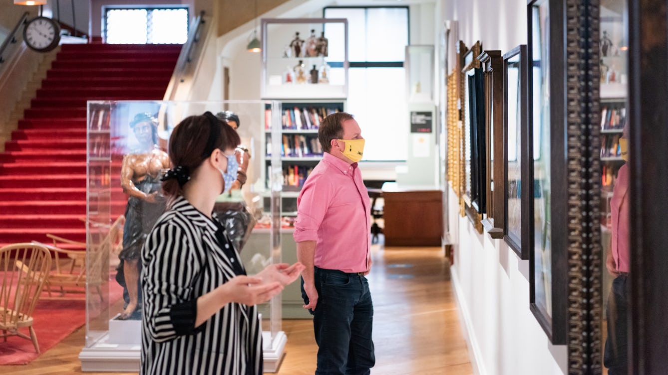 Photograph of a museum gallery space with free standing book shelves, wooden chairs, framed artworks on the wall and a red staircase in the distance. Standing in the centre of the image is a man in a pink shirt wearing a yellow face covering. He is looking at a framed artwork on the wall. Next to him is a woman in a stripy jacket, also wearing a face covering. She is also looking at the artworks and has her arms raised as if in conversation.