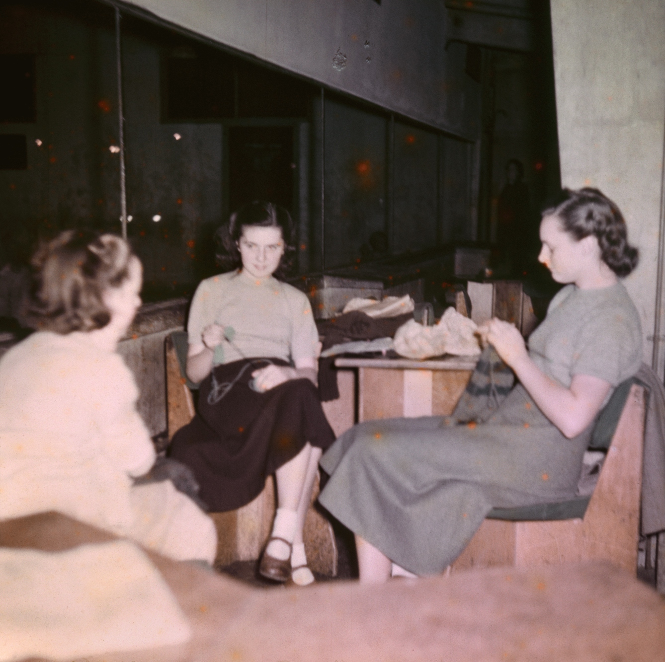 Photograph from the mid-20th Century Peckham Pioneer Centre, showing three women seated at a table, knitting. The building in which they sit has concrete columns and large windows.