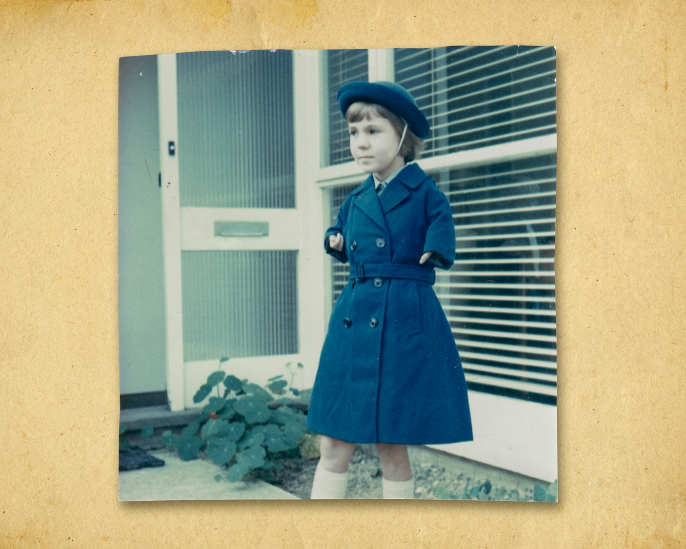 Photograph of a colour photographic print roughly cut around the edges, resting on a brown paper textured background. The print shows a small young girl in a long blue coat and matching blue hat. She is standing outside a house with a large front window and glass panelled front door which is open. On the ground are the leaves of a plant. The girl is looking off to the left. Her arms are short as a result of her mother being prescribed Thalidomide during her pregnancy. 