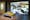 一个画廊装置的照片，展示了一张有黄色靠垫的长凳，面对着一个大的视频投影。投影显示了麦当劳餐厅的内部，从高处向下看，餐厅的地板开始被水淹没。