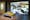 Photograph of a gallery installation showing a bench with yellow cushions facing a large video projection. The projection shows the inside of a McDonalds restaurant looking down from a high viewpoint in which the restaurant floor is beginning to flood with water.